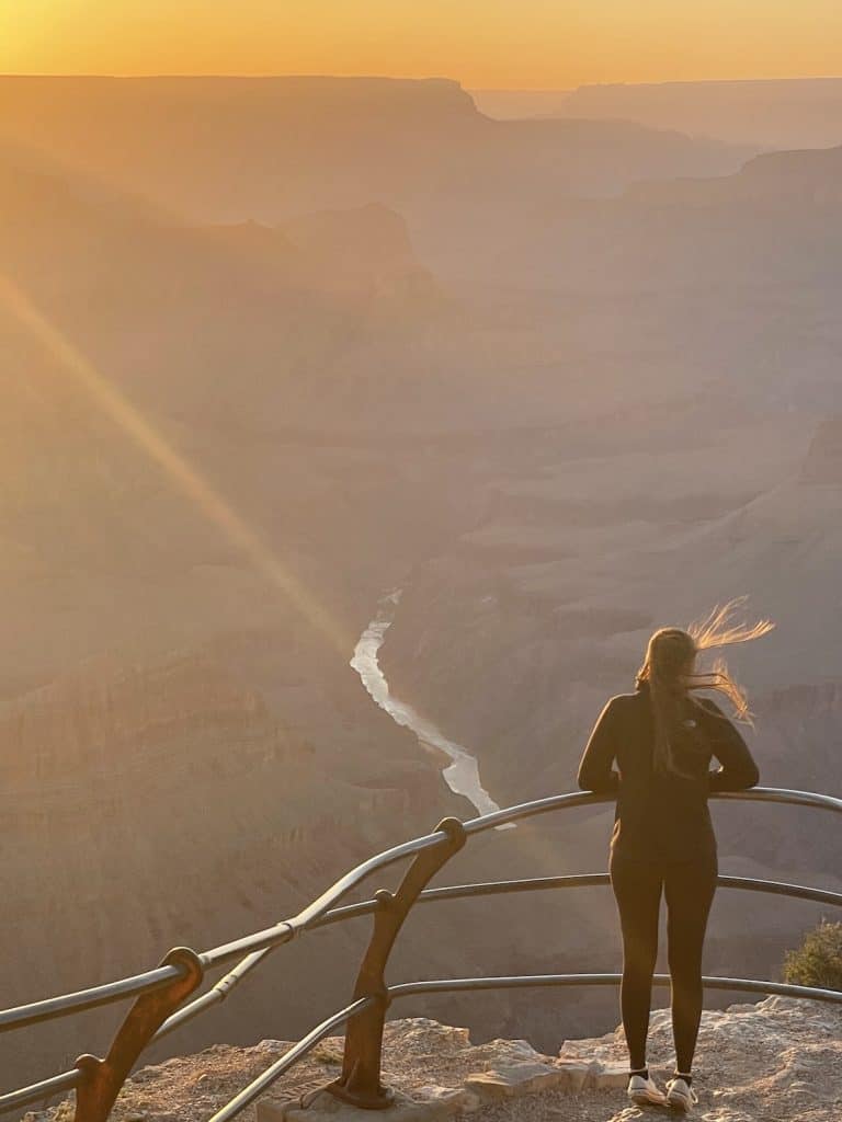 girl-on-balcony-ledge-looking-over-sweeping-grand-canyon-with-colorado-river-straight-ahead-sunset-washes-out-the-colors-her-pony-tail-blows-in-the-wind-what-to-do-with-one-day-grand-canyon-national-park-south-rim-northern-arizona-mohave-point