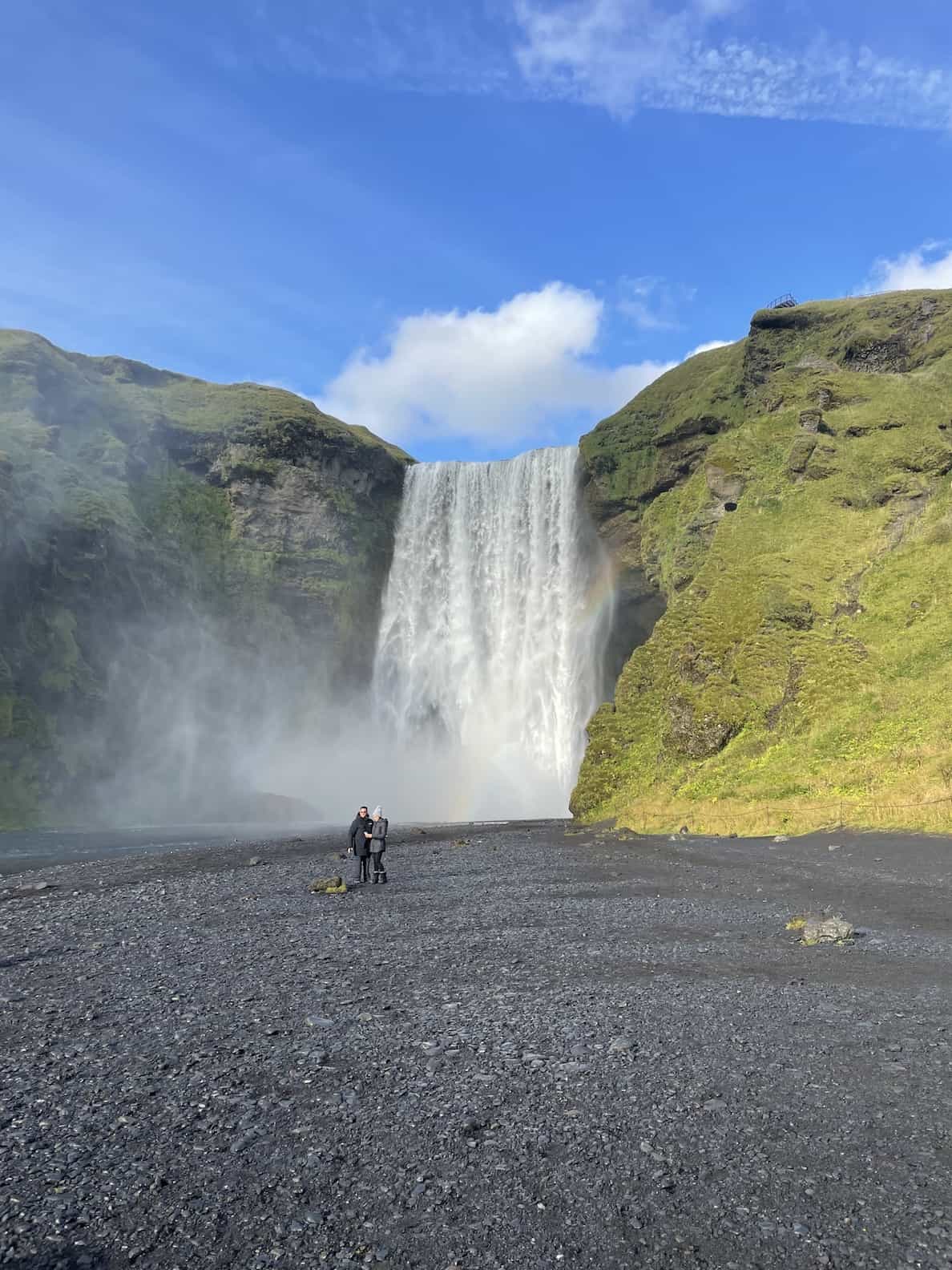 a-couple-standing-in-front-of-the-powerful-197-foot-by-85-foot-freefalling-Skogafoss-waterfall-on-a-sunny-day-with-rainbow