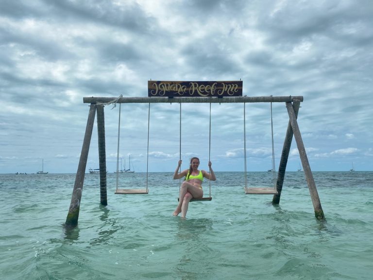 Woman swinging on wooden overwater swing surrounded by the bright blue Caribbean Sea with sailboats in background. Iguana Reef Inn, Caye Caulker, Belize