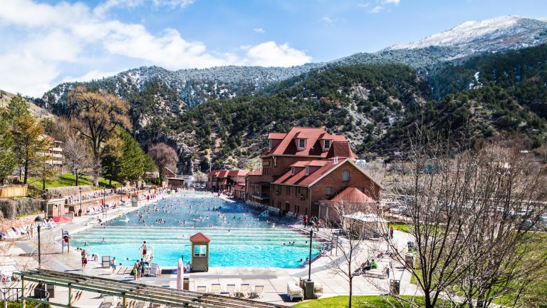 Large hot spring pool next to historic brick building under large, snow covered mountains on sunny winter day. Glenwood Springs Hot Springs, Glenwood Springs, Colorado.