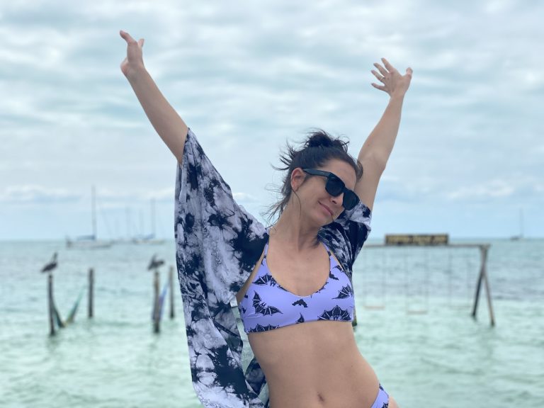 Smiling girl waving her hands in the air wearing purple bikini and tie-dye beach cover up. In background is bright green waters of the Caribbean Sea. Caye Caulker, Belize.