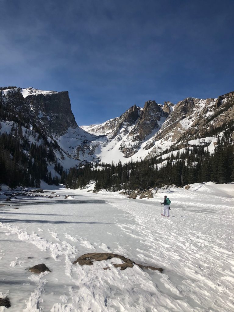 Woman snowshoeing across frozen, snow covered lake with huge rocky mountain peaks in background on sunny winter day. Snowshoeing Dream Lake, Rocky Mountain National Park, near Estes Park, Colorado.