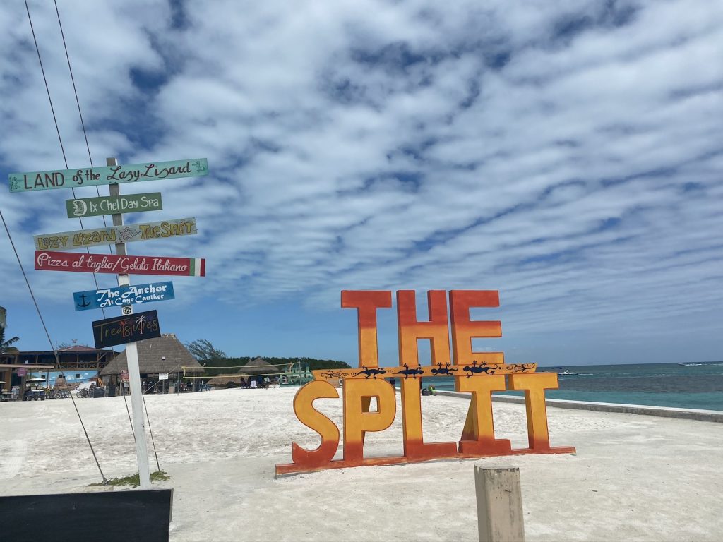 large orange sign reading "The split" on beach with beach bars and Caribbean Sea in background.