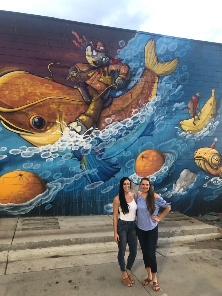 Two woman smiling in front of large, colorful mural at Denver Blue Moon Brewery in RiNo Neighborhood. Blue Moon Brewery, River North (RiNo), Denver, Colorado.