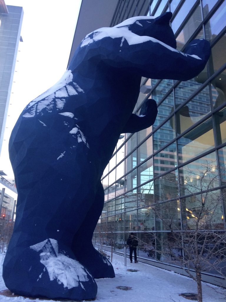 Big blue bear peeking into the huge windows of the Colorado Convention Center with small person standing below the bear, bear covered in snow during winter. Big Blue Bear, Colorado Convention Center, Denver, Colorado.