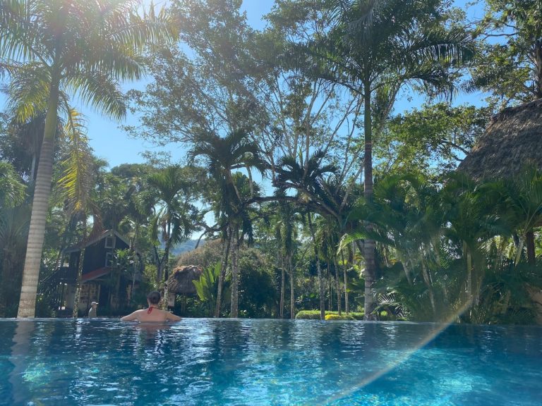 Woman with back to camera overlooking lush green jungle and thatch-roof resort buildings from beautiful infinity pool. Infiniti pool at at Lodge At Chaa Creek, San Ignacio, Belize.