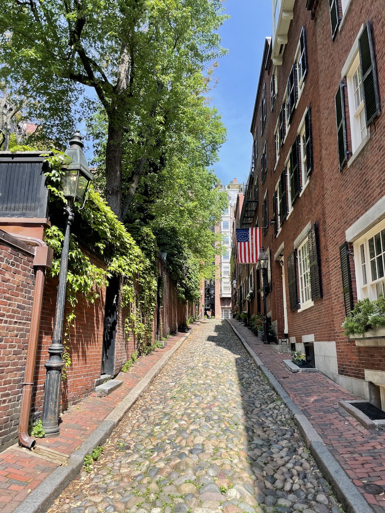 Small alleyway with brick sidewalks, cobblestone street, lined with brick buildings with shingles and American Flag on sunny summer day. Acorn Street, Beacon Hill, Boston, Massachusetts, New England.