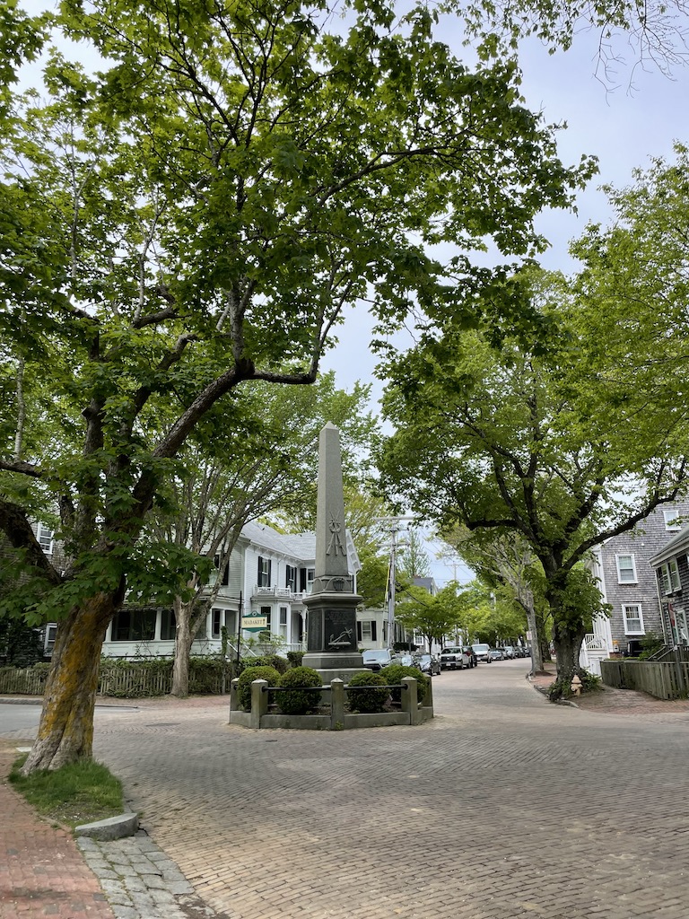 Cobblestone street surrounded by historic homes and small roundabout with historic monument in center. Road is lined with historic homes, brick sidewalks, and mature trees. Nantucket, Cape Cod, Massachusetts.