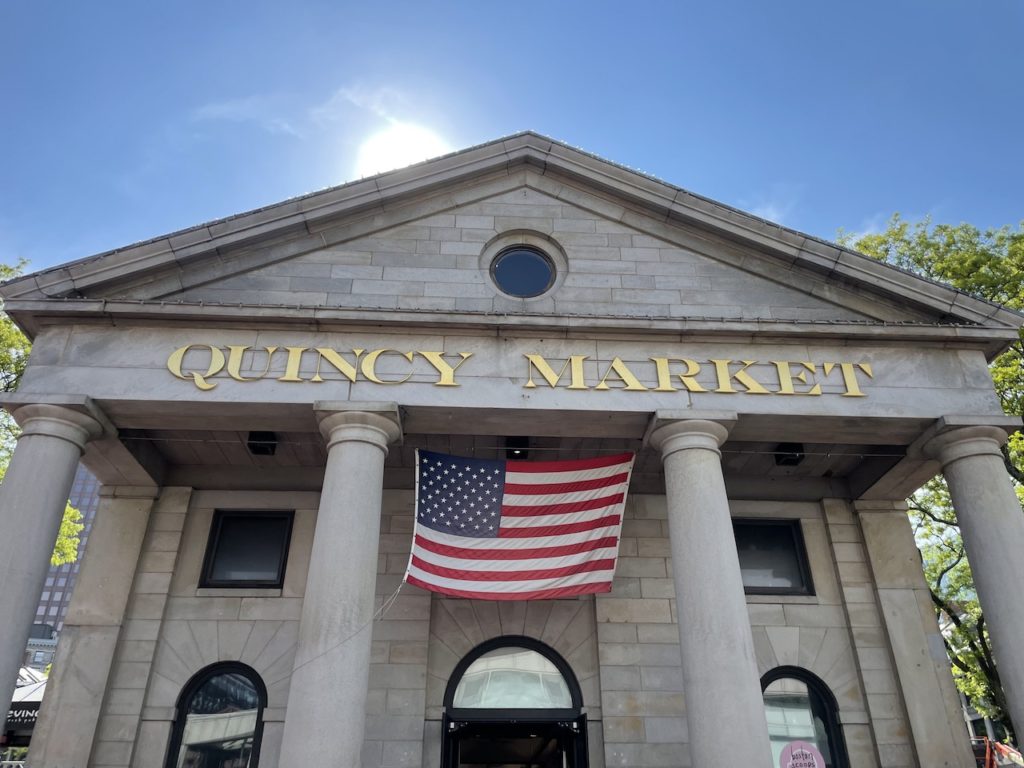 Two-story granite building with large American flag and gold sign reading "Quincy Market". Quincy Market, Faneuil Hall Marketplace, Boston, Massachusetts, New England.