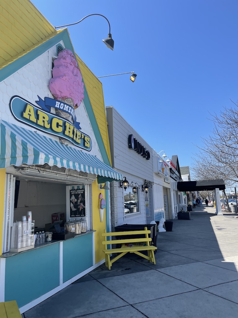 Colorful ice cream shop with pink/yellow/teal ice cream sign reading "Archies Homemade" colorful building sits along side row of storefronts on sunny summer day. Shopping on Rehoboth Avenue in Rehoboth Beach, Delaware.