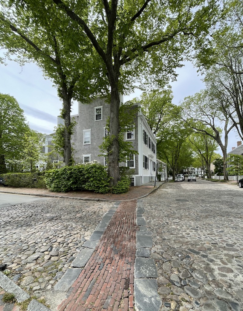 Old cobblestone and brick road leading to shingled historic home surrounded by large, green trees on gloomy early summer day. Historic Nantucket, Cape Cod, Massachusetts, New England.