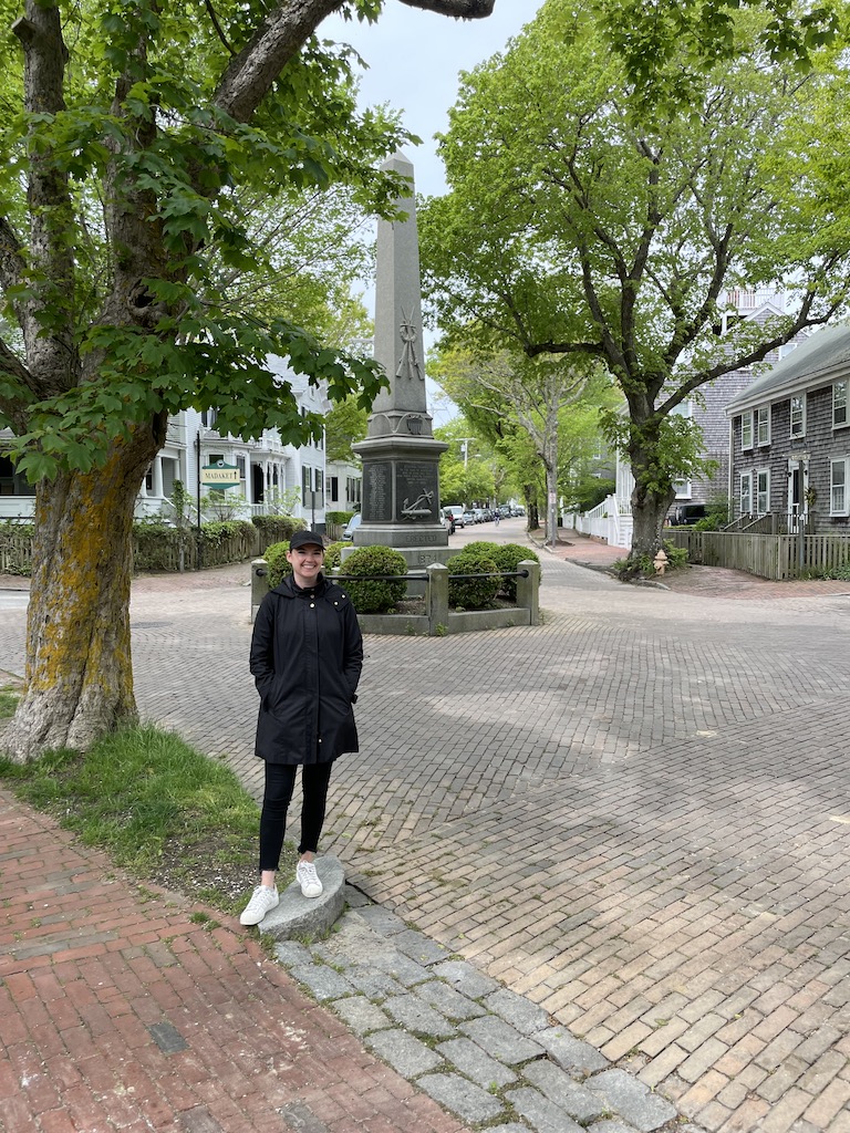 Woman standing on cobblestone street surrounded by historic homes and small roundabout with historic monument in center. Road is lined with historic homes, brick sidewalks, and mature trees. Nantucket, Cape Cod, Massachusetts, New England.
