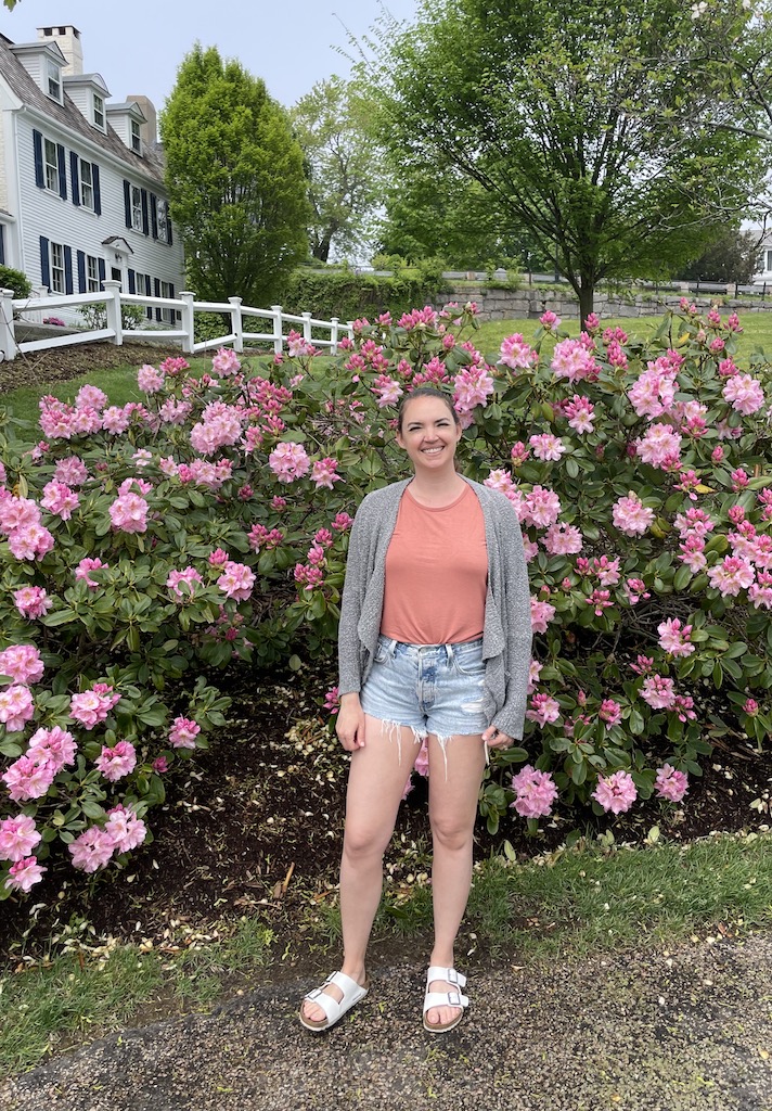 Woman standing in front of large rose bush with stately white mansion in background. Brewster Park, Plymouth, Massachusetts.