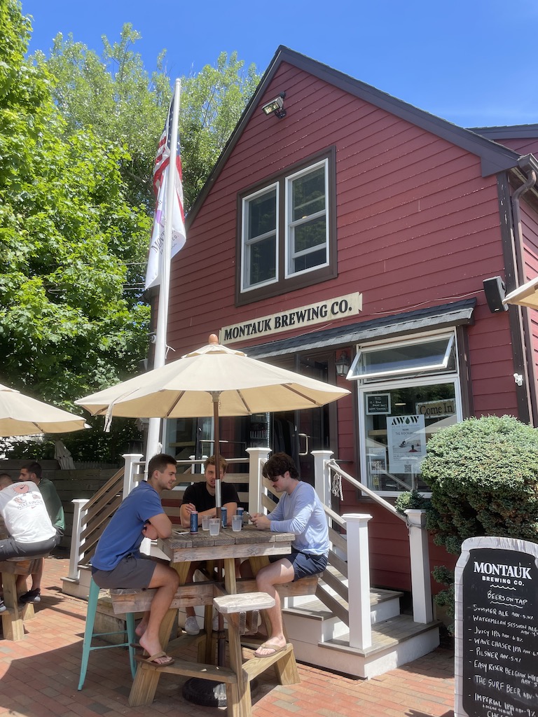 Red barn building with outdoor patio seating in front full of people under umbrellas on sunny summer day. Montauk Brewery, Montauk, New York, The Hamptons.
