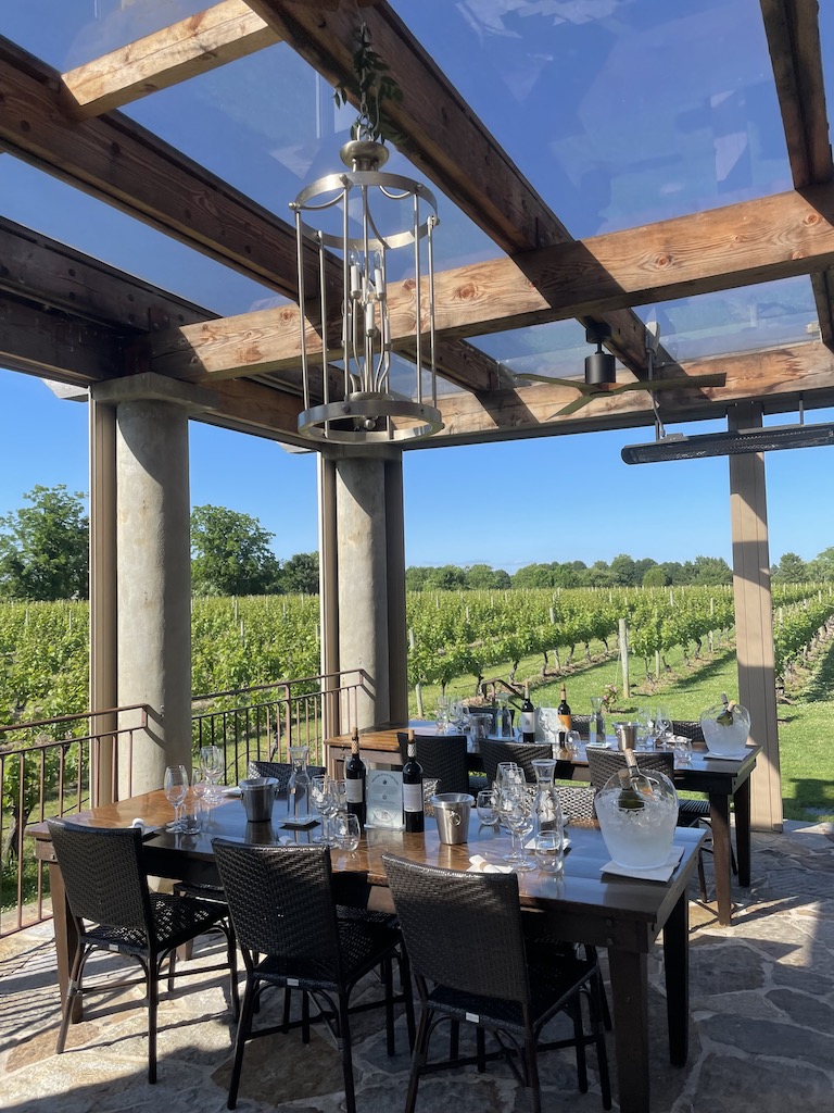Luxury table set up on outdoor patio covered in terrace with chandelier, view overlooks surrounding lush green vineyard on cloudless summer day. Wolffer Estate Vineyards, wine tasting, Sagaponack, New York, The Hamptons.