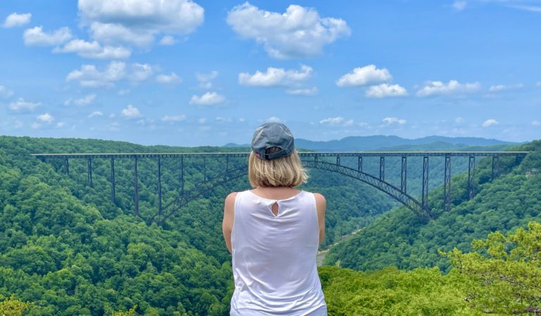 Woman looking out at huge steel bridge spanning bright green gorge during summer. New River Gorge Bridge, New River Gorge National Park, West Virginia.