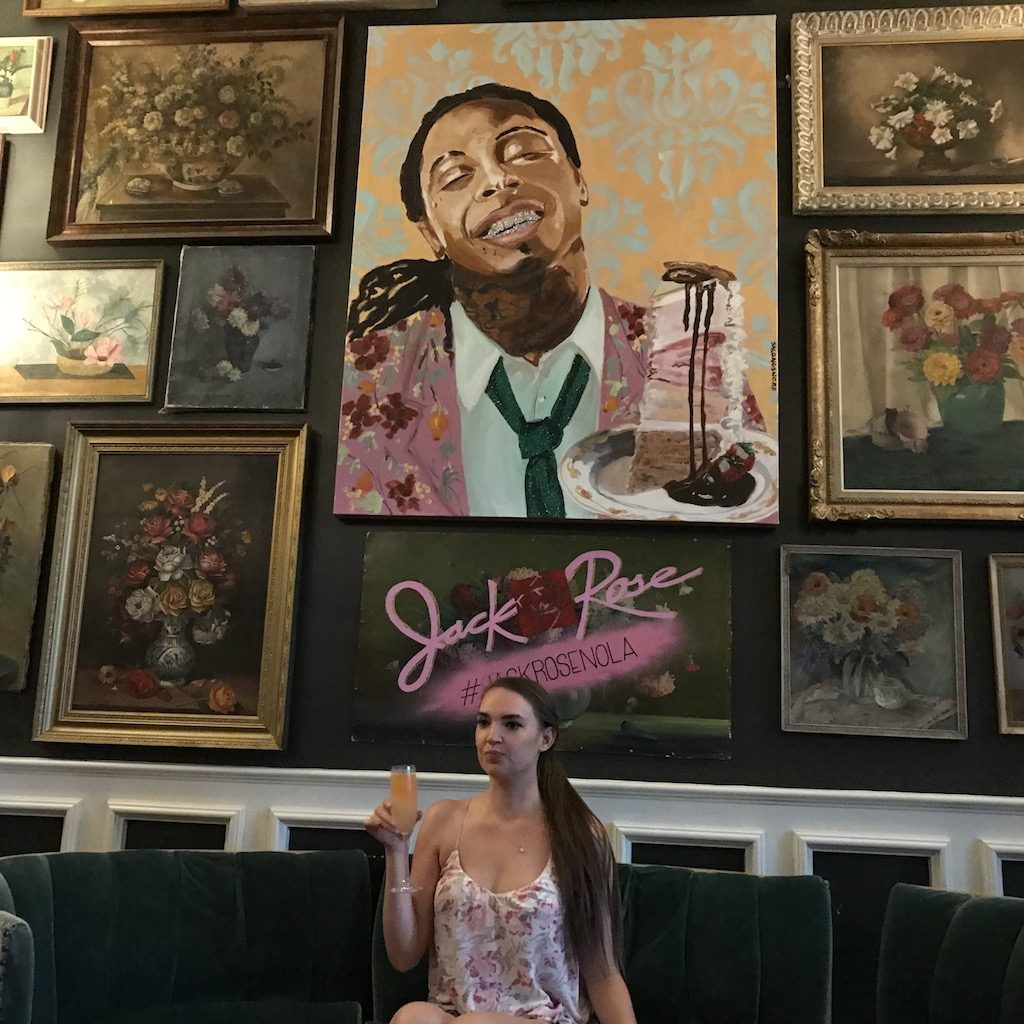Woman sitting on plush green couch with a glass of Champagne in front of wall covered in fancy paintings with a large painting of Lil Wayne with a piece of cake in center. Pontchartrain Hotel, New Orleans, Louisiana.