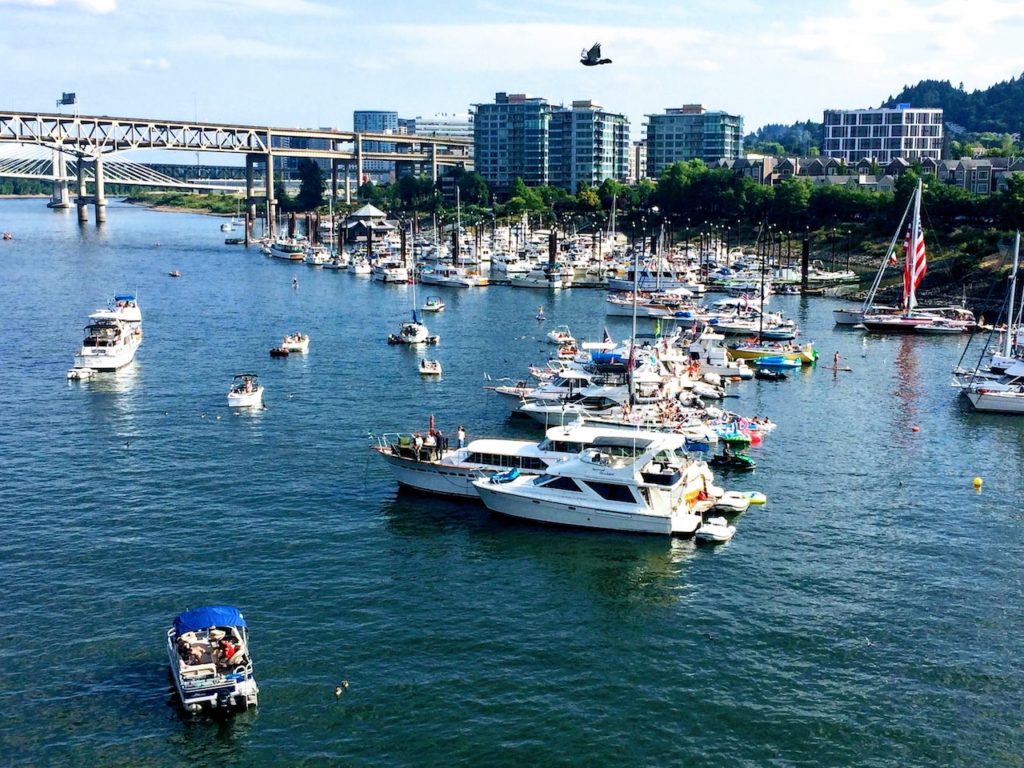 Recreational boats and yachts lined up in Willamette River near harbor full of boats with bridges and a few high rise buildings in background. Sunny, summer day in downtown Portland, Oregon.