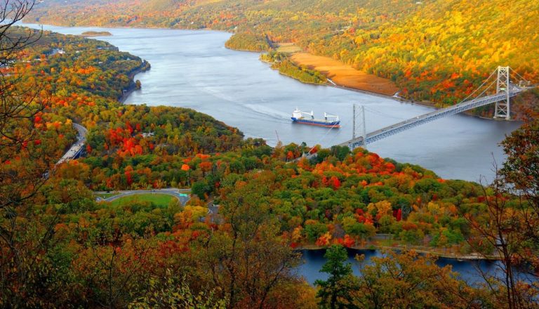 Ariel view of wide, calm Hudson River with large bridge spanning river and large boat making its way down river. River is surrounded by colorful mountainsides of red, orange, yellow, and green during fall. Bear Mountain State Park, Hudson Valley, Upstate, New York.