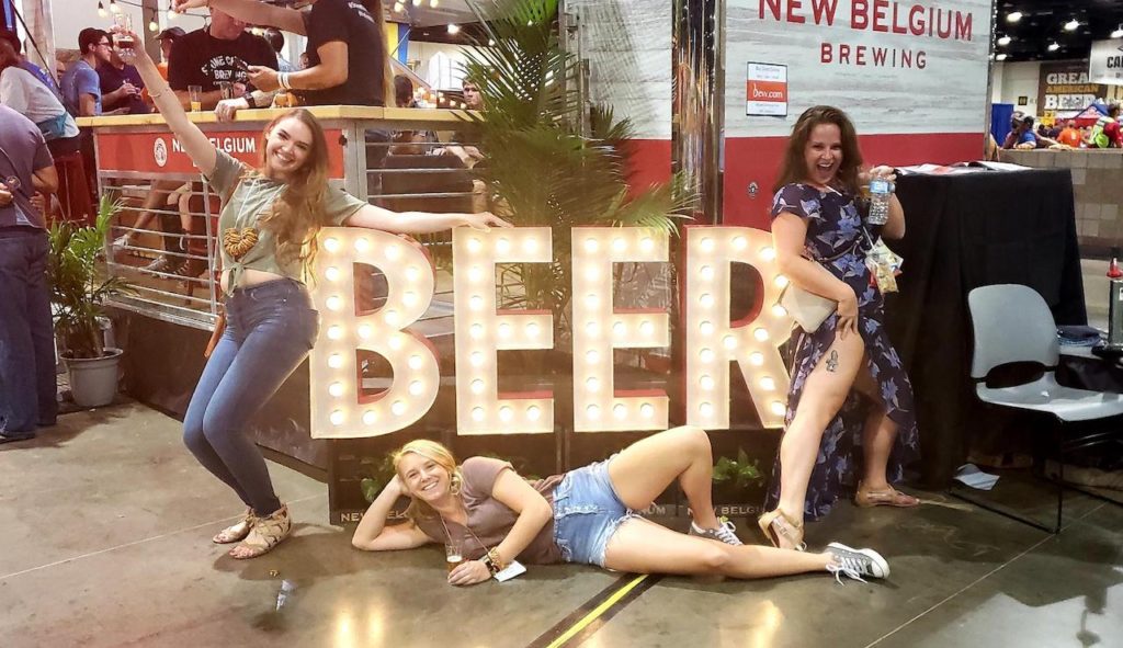 Three woman laughing and posing with large, lit up sign reading "BEER". Great American Beer Festival, Denver Convention Center, Downtown Denver, Colorado. Things to do in Denver during fall, fall activities Colorado