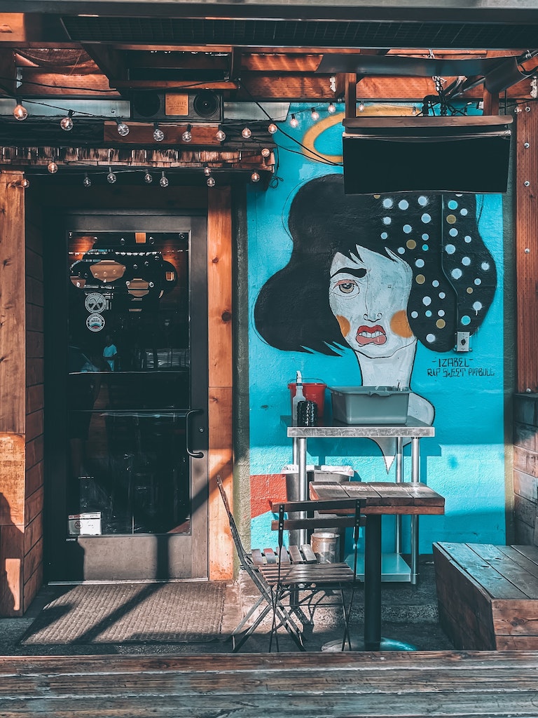 Wooden building, business entrance with unlit string lights an bright blue mural of abstract woman with one eye and polka dot hair. Portland art district, Portland Oregon.
