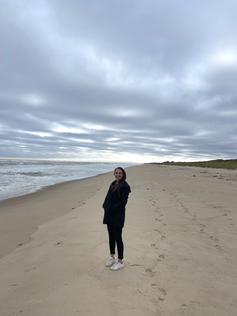 Woman in rain jacket standing on white sand beach with Atlantic Ocean in background on grey, cold day. Atlantic Avenue Beach, Amagansett, New York, The Hamptons.
