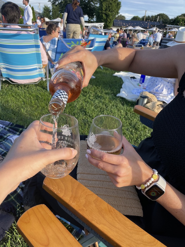 Woman pouring wine into wine glass on grassy lawn full of people in lawn chairs and picnic blankets with live music. The Wine Stand at Wolffer Estate Vineyard, Sagaponack, New York, The Hamptons.