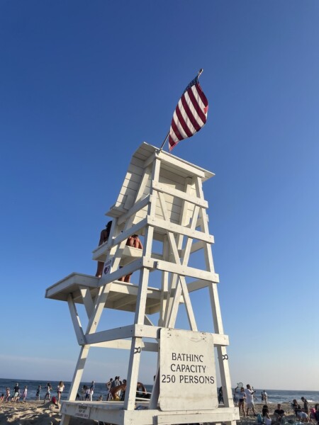White wooden lifeguard tower with American flag overlooking beach full of people and Atlantic Ocean on cloudless summer day. Main Beach, East Hampton, New York, The Hamptons.