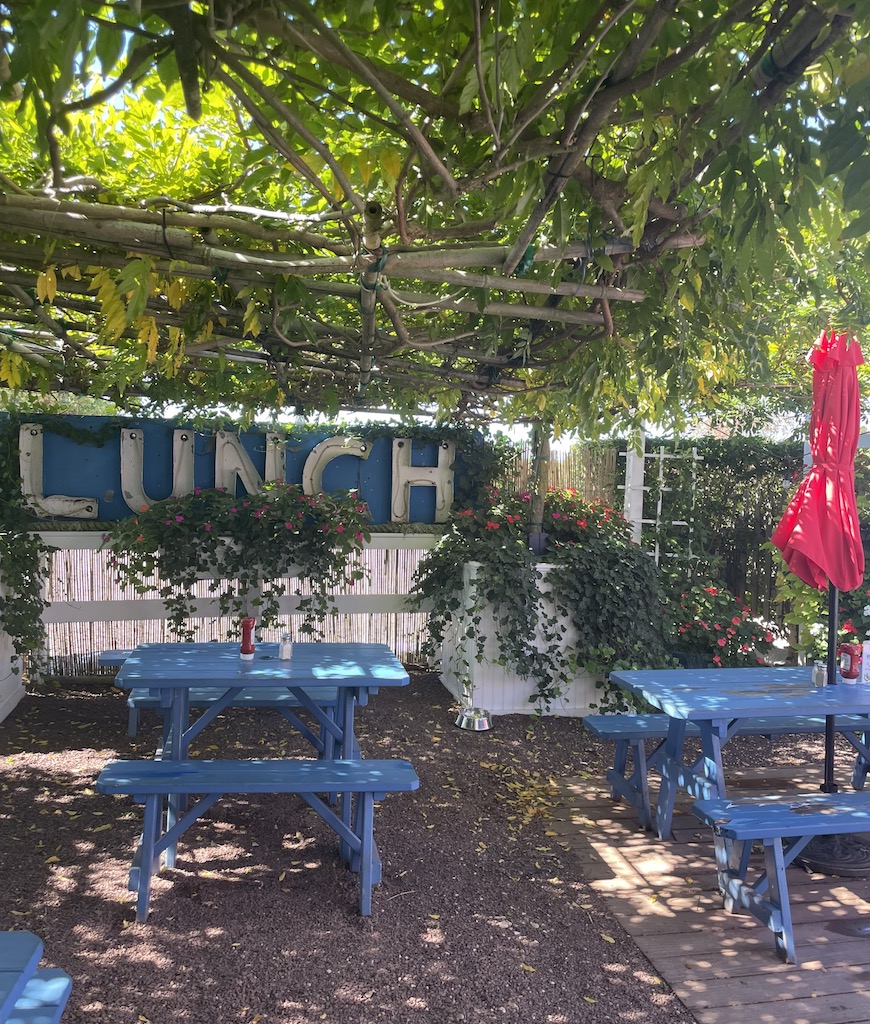 Outdoor garden courtyard covered in green, live awning of plants, surrounded by flowers, blue wooden picnic tables sit under shade in front of large antique sign reading "LUNCH". Lobster Roll AKA Lunch, Amagansett, New York, The Hamptons.