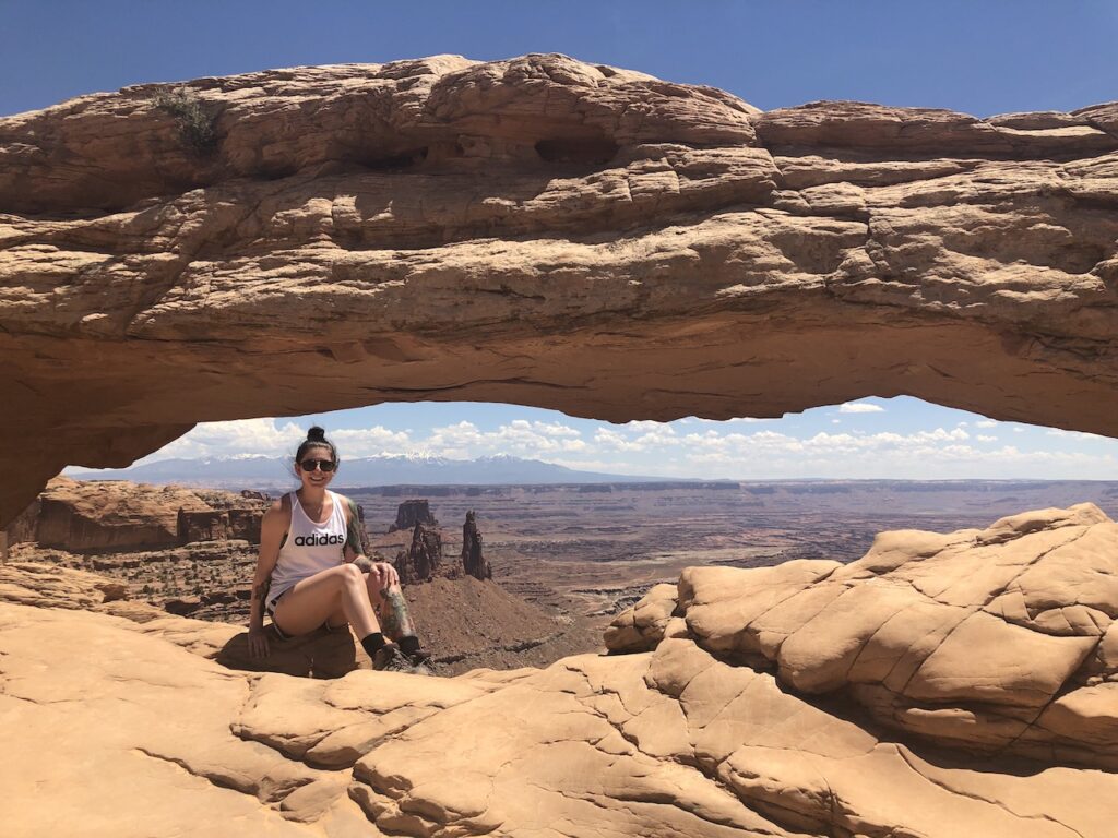 Woman sitting under low, long red rock arch with dramatic desert canyon views in background on warm summer day. Mesa Arch, Canyonlands National Park, Utah.