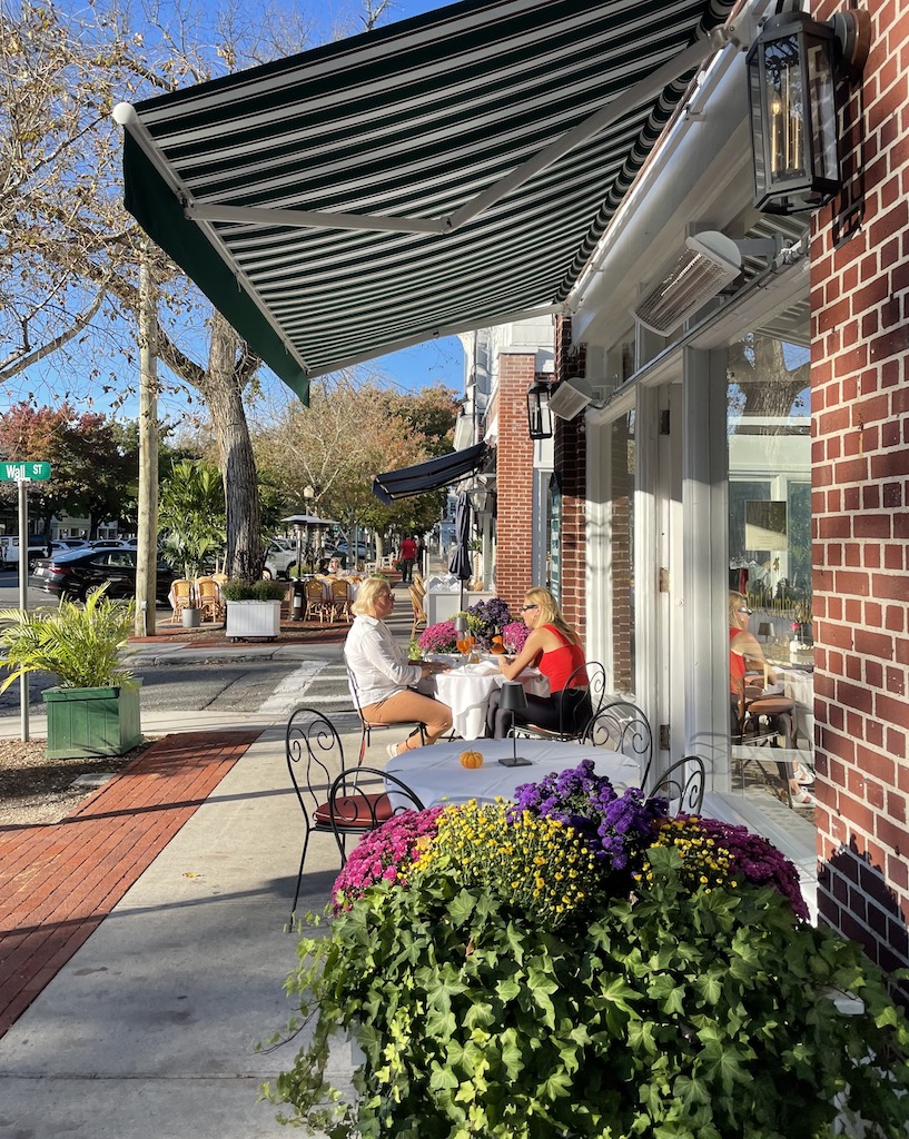 Two woman at bistro table by large pot of flowers enjoying outdoor sidewalk seating at a restaurant on Main Street in Southampton. Southampton, New York, The Hamptons.
