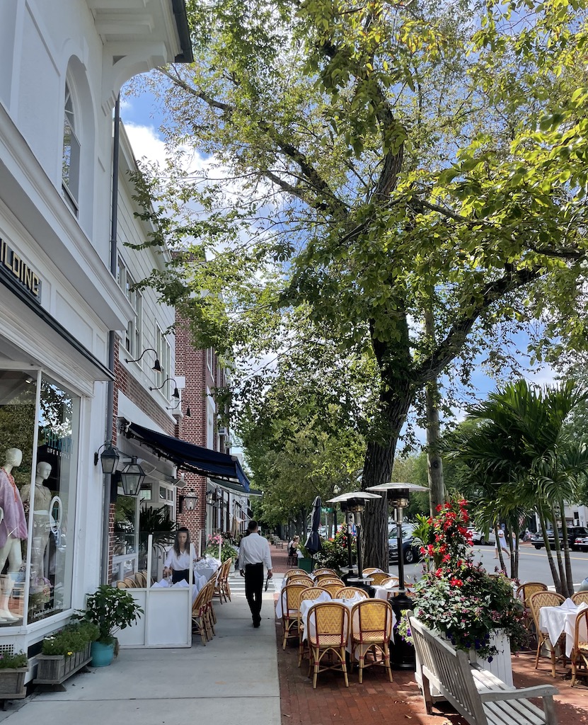 Sidewalk cafe seating framed by buildings and large, mature trees during a sunny summer day. Main Street, Southampton, New York, The Hamptons.