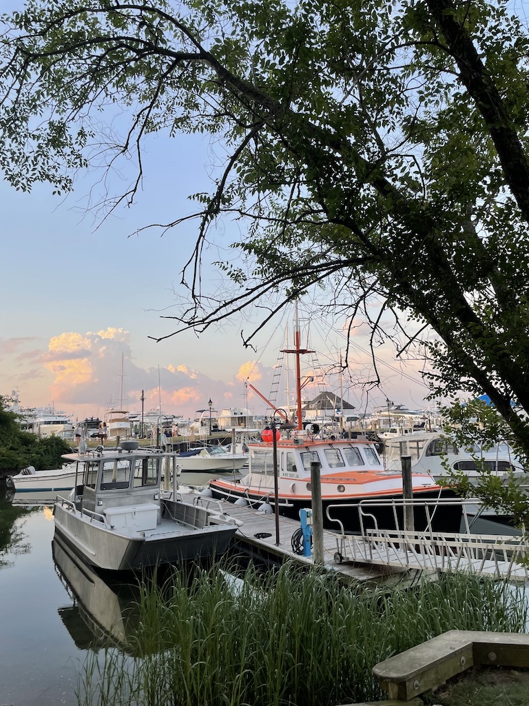 Yacht yard filled with boats including colorful orange and black boat. In front of yacht yard are large trees, in background sunset. Sag Harbor Yacht Yard, Sag Harbor, New York, The Hamptons.