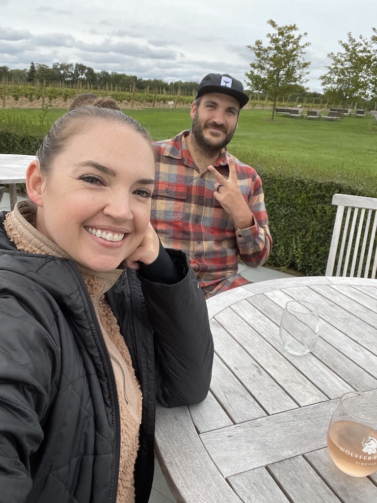 Couple smiling at wooden table with stemless wine glasses on grey cold day, background is a vineyard and grass lawn. Wine Stand at Wolffer Estate Vineyard, Sagaponack, New York, The Hamptons.