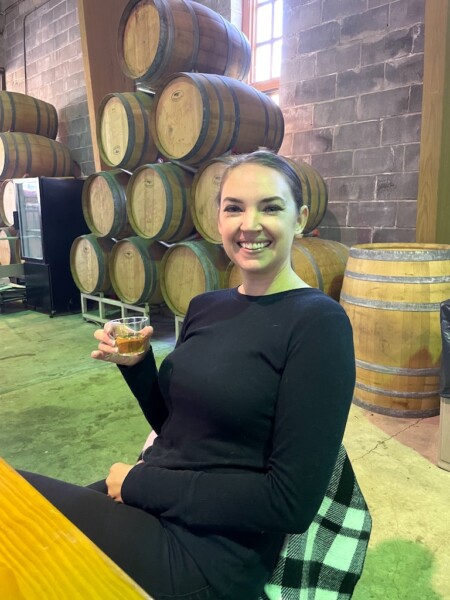 Woman in indoor tasting room holding glass of wine with large barrels of wine stacked against wall in background of warehouse-like room. Duck Walk Vineyards, Water Mill, New York, The Hamptons.