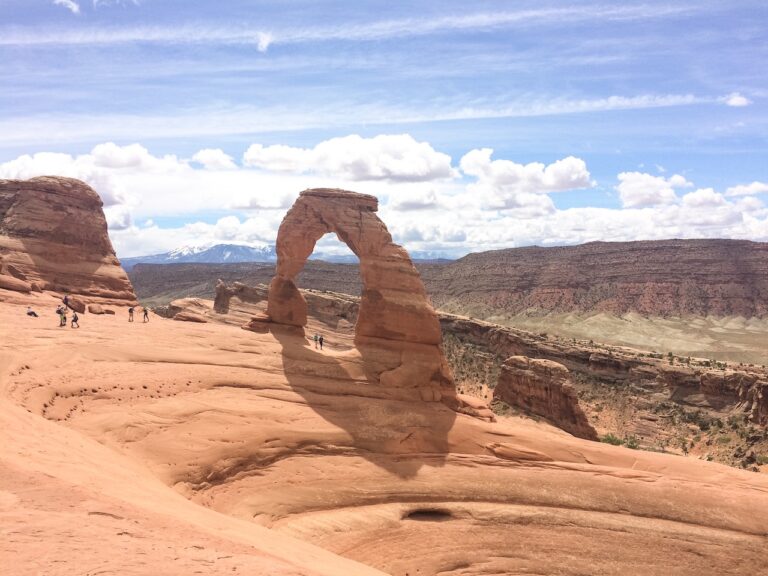 Large, freestanding red rock arch rising out of solid red rock surrounded by desert landscape and snow covered mountains. Hikers at Delicate Arch, Arches National Park, Moab, Utah.