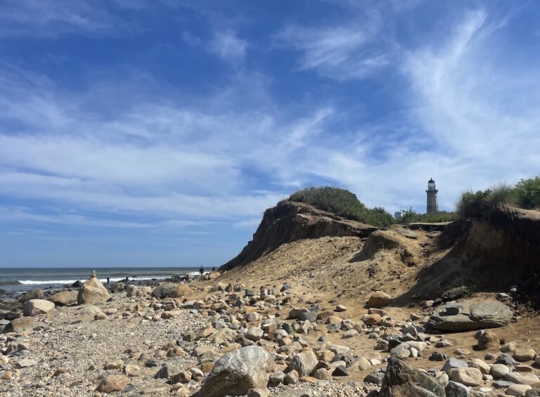 Sandy beach with large rocks and small beach cliffs overlooking Atlantic Ocean, lighthouse in distance. Montauk Point Lighthouse, Montauk Point State Park, Montauk, New York, The Hamptons.