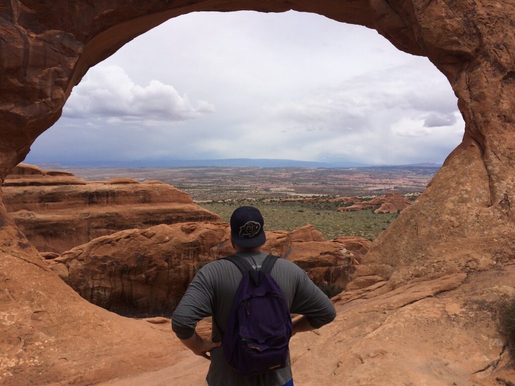 Man standing in front of red rock arch looking out at views of the surrounding valley on the other side. Partition Arch, Arches National Park, Moab, Utah.