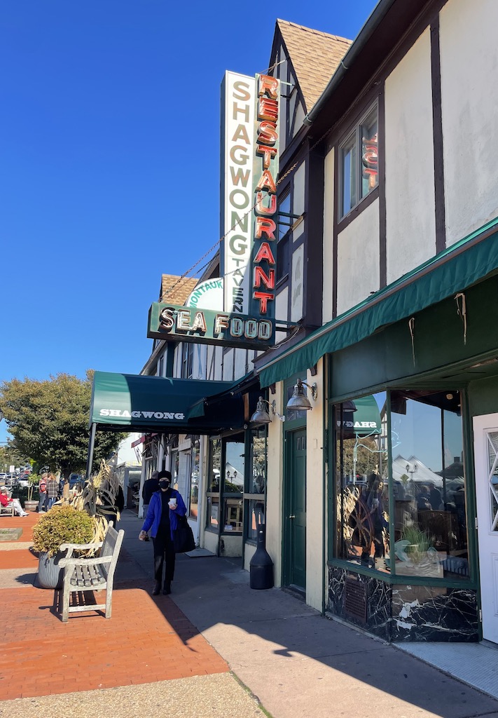 Building in downtown shopping area with pedestrians walking on sidewalk, sign on building reads "Shagwong Restaurant Sea Food". Montauk, New York, The Hamptons.