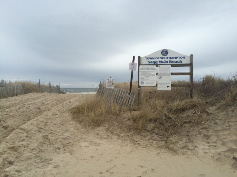 Sandy shore on grey winter day with Atlantic Ocean in background, beach sign reads "Town of Southampton Sagg Main Beach". The Hamptons, Sagaponack, New York