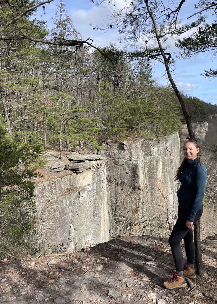 Woman hiking with rocky sheer cliffs covered in pine trees at top. Endless Wall Trail, New River Gorge National Park, West Virginia.