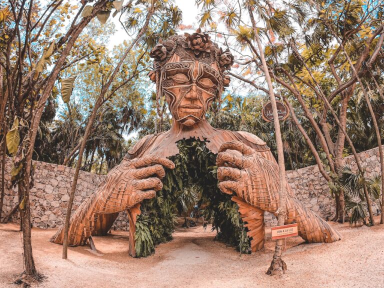 Wooden statue of person in with elaborate geometrical details, person is opening chest which is covered in plant greenery and serves as walkway/ entry way to hotel. Statue is surrounded by palm trees and plants, gravel road. Coming To Light Sculpture, Tulum, Mexico.