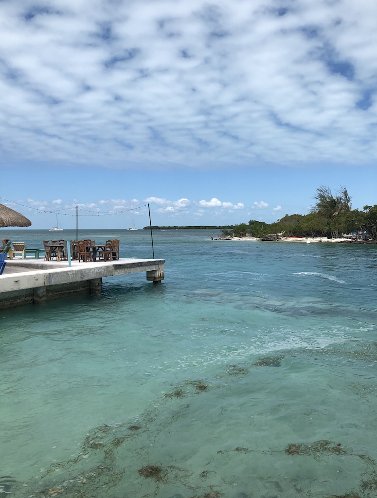 Clear blue water surrounding beach bar dock over water with lush, green island in background on sunny day. The Split, Caye Caulker, Belize.