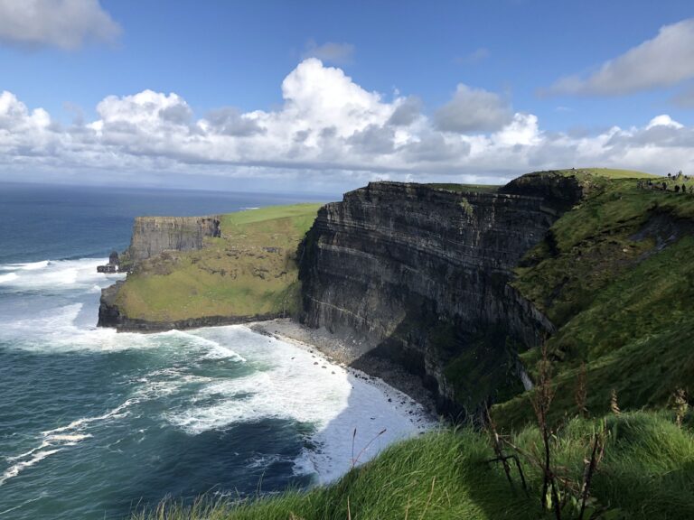 Sheer stone cliffs covered in green grass over bright blue water of the Atlantic Ocean on sunny, clear day. Cliffs of Moher, Ireland, Europe.
