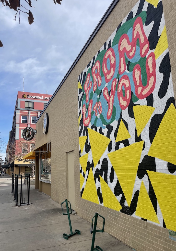 Tan brick building with colorful mural, yellow, with geometric shapes and pink/ red text reading "From U To You" in background is more buildings and sunny skies. Downtown Colorado Springs, Colorado.