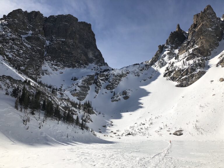 Jagged, rocky mountains covered in snow, in foreground is frozen snow-covered lake with tiny hiker in distance. Emerald Lake, Rocky Mountain National Park, Colorado.