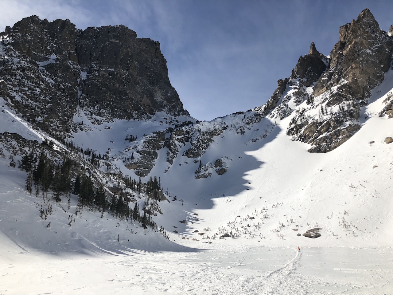 Jagged, rocky mountains covered in snow, in foreground is frozen snow-covered lake with tiny hiker in distance. Emerald Lake, Rocky Mountain National Park, Colorado.