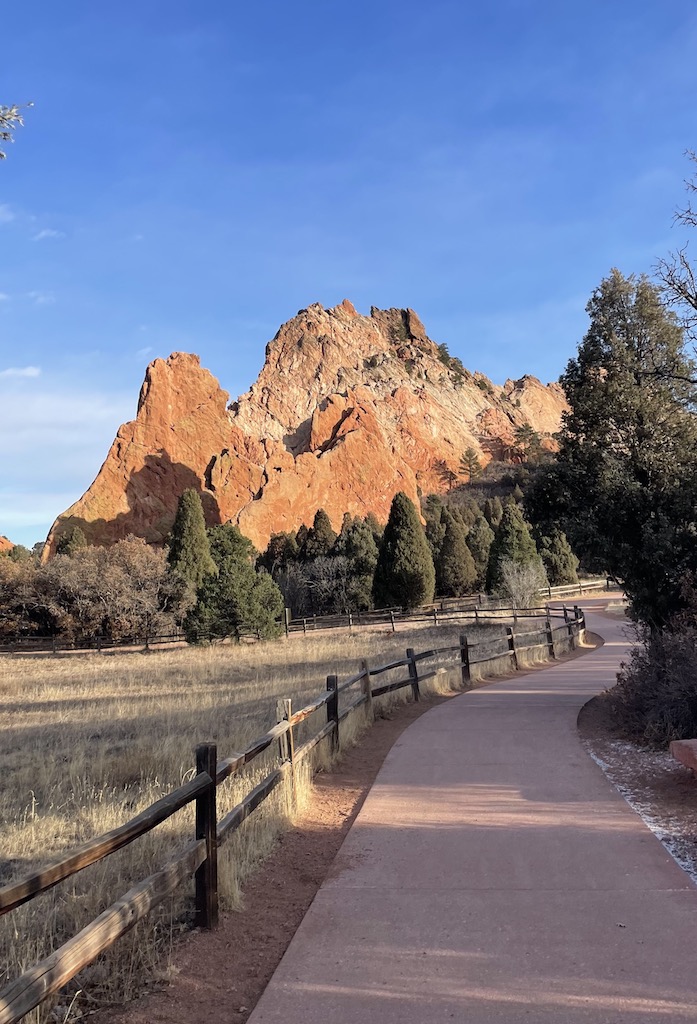 Huge red rocks jutting up from earth with paved path winding towards unique landscape surrounded by trees on sunny day. Garden of the Gods, Colorado Springs, Colorado.