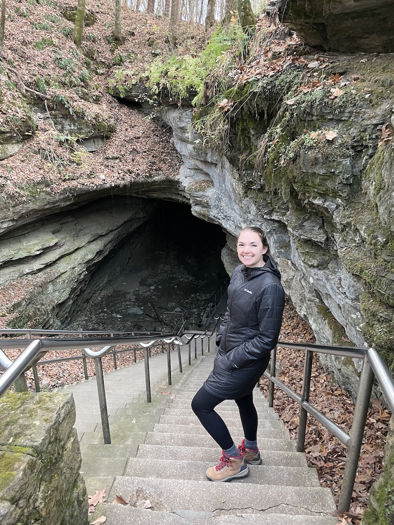 Woman standing at mouth of rocky cave covered in greenery. Mammoth Cave National Park, Kentucky.