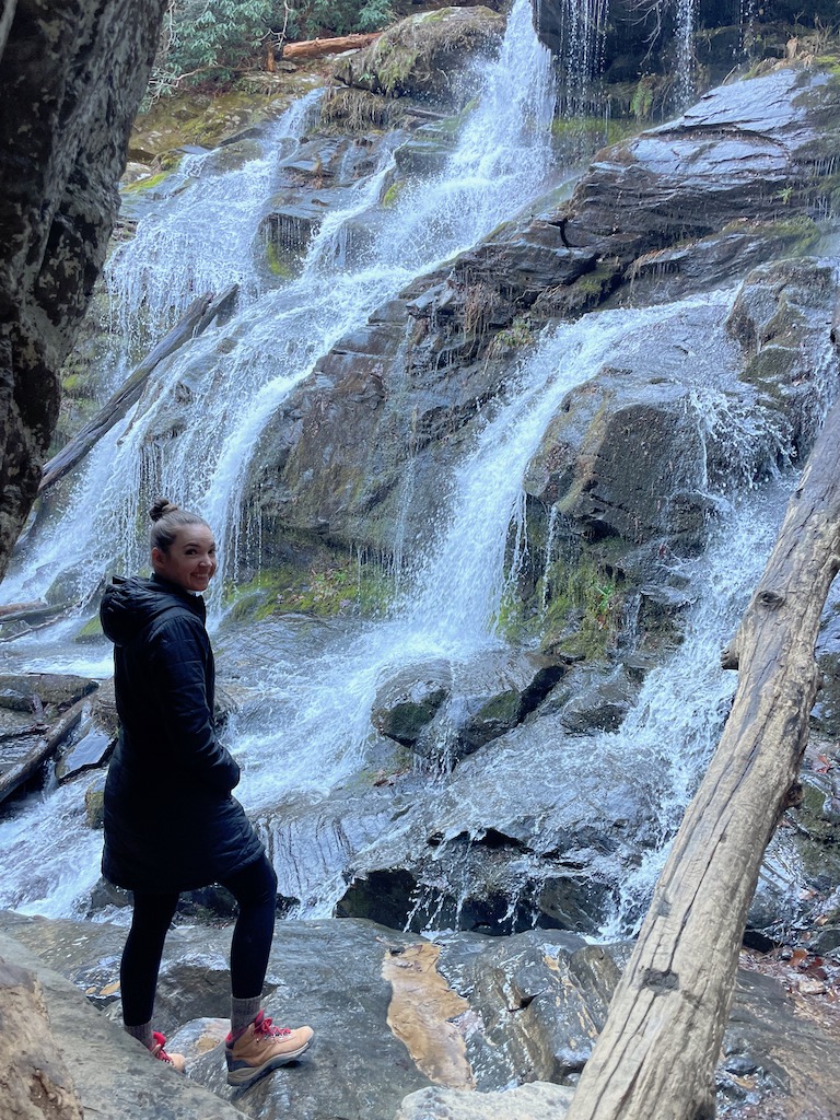 Woman in winter jacket smiling at camera with huge rocky, waterfall in background. Pisgah National Forest, Asheville, North Carolina.